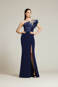 A woman modeling a floor-length navy blue gown with a one-shoulder ruffled neckline, a thigh-high slit, and a silver-toned embellished motif on the bodice, photographed against a plain light beige studio background.