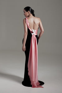 A model is shown from the back wearing a floor-length evening gown with a black body and a draped pink fabric panel across the back and shoulder, accented with white floral details at the waist, against a neutral gray studio background.