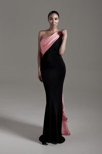 A woman modeling a floor-length one-shoulder evening gown with a black body and a draped pink satin overlay, photographed against a plain gray studio background.