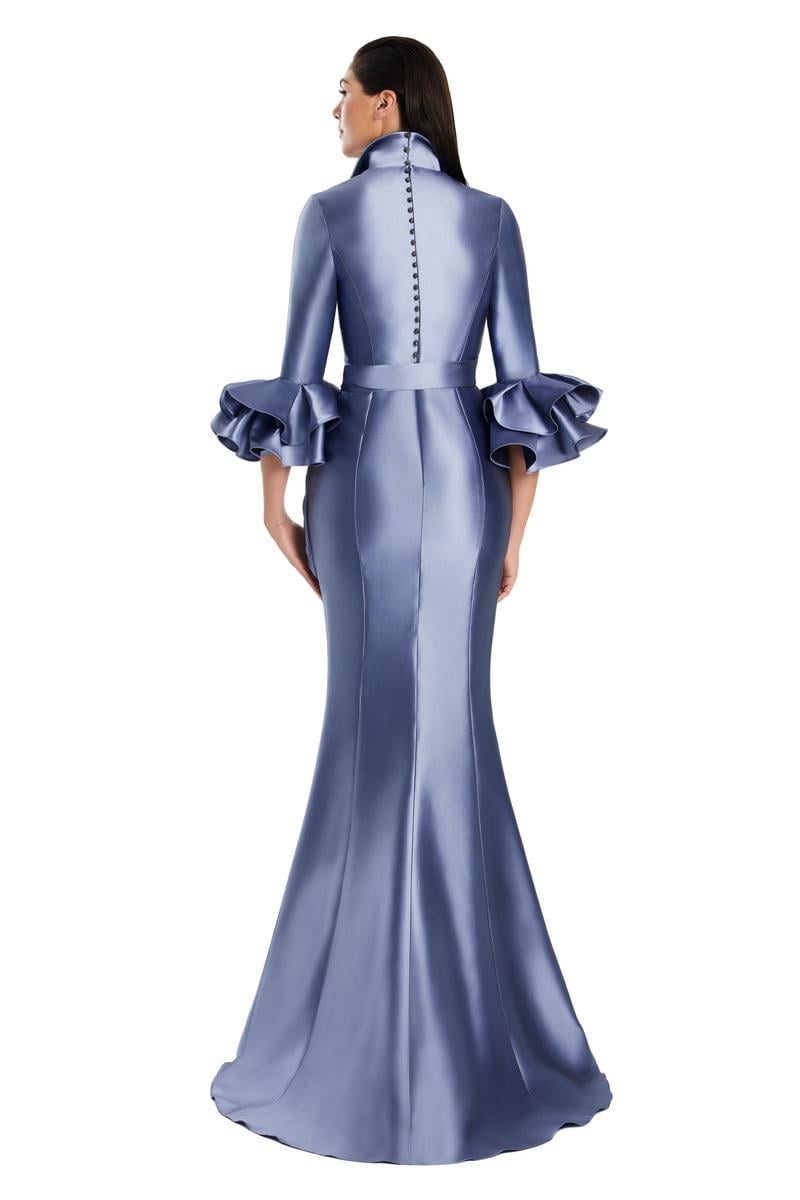 Model wearing Alexander by Daymor 3087S25 gown with mermaid skirt and high-neck jacket featuring dramatic ruffle sleeves and waist sash.