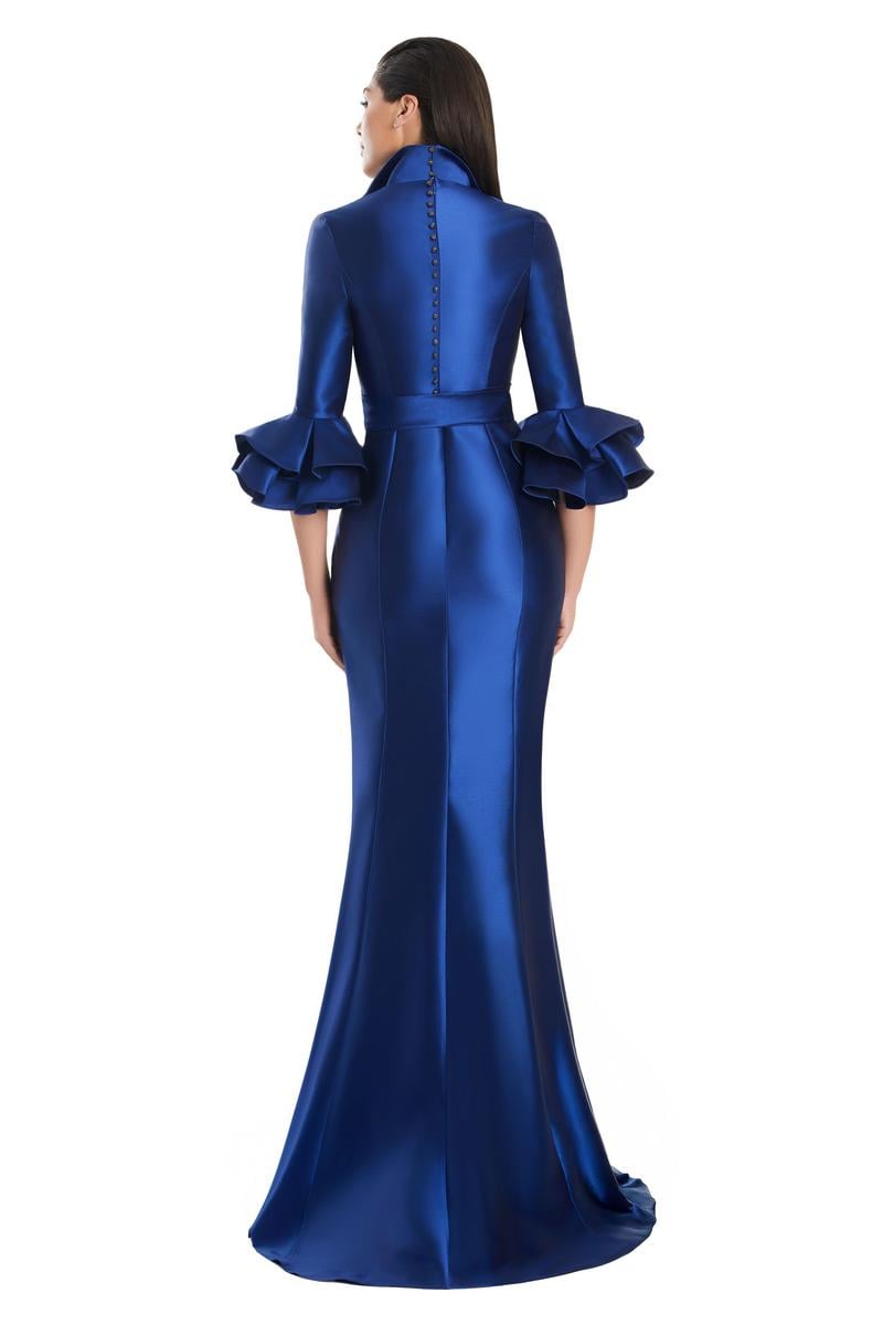 Model wearing Alexander by Daymor 3087S25 gown with mermaid skirt and high-neck jacket featuring dramatic ruffle sleeves and waist sash.