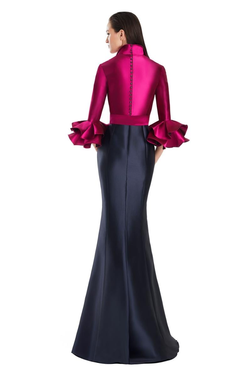 Model wearing Alexander by Daymor 3087S25 gown with mermaid skirt and high-neck jacket featuring dramatic ruffle sleeves and waist sash.