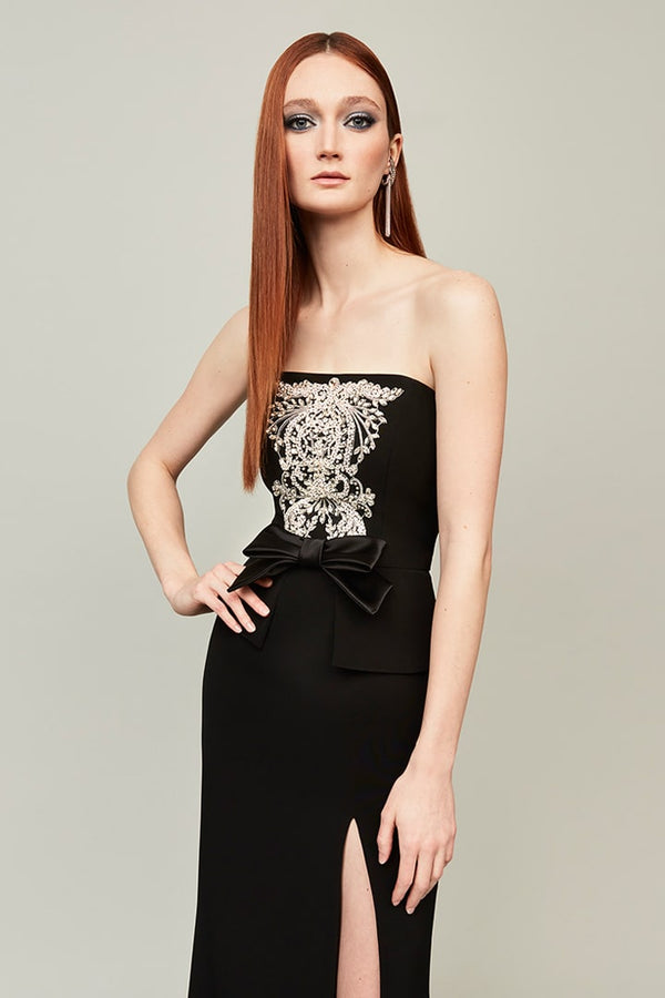 A woman modeling a strapless black gown with a large white ornate brocade pattern across the bodice, a black bow at the waist, and a thigh-high slit, against a solid light gray background.