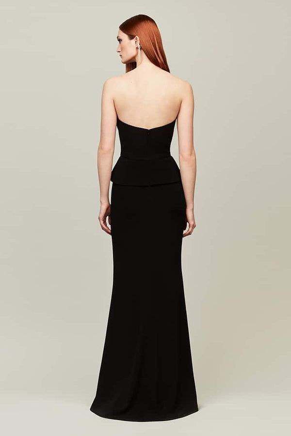 A woman with red hair is shown from the back wearing a strapless black gown with a structured corset-style bodice and peplum, photographed against a plain light gray studio background.