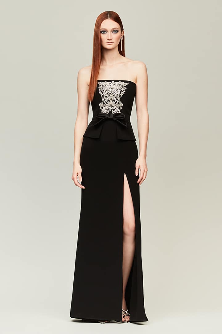 A woman modeling a strapless black evening gown with a high thigh slit and a large silver floral applique on the bodice, photographed against a plain light gray studio background.