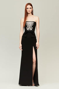 A woman modeling a strapless black evening gown with a high thigh slit and a large silver floral applique on the bodice, photographed against a plain light gray studio background.