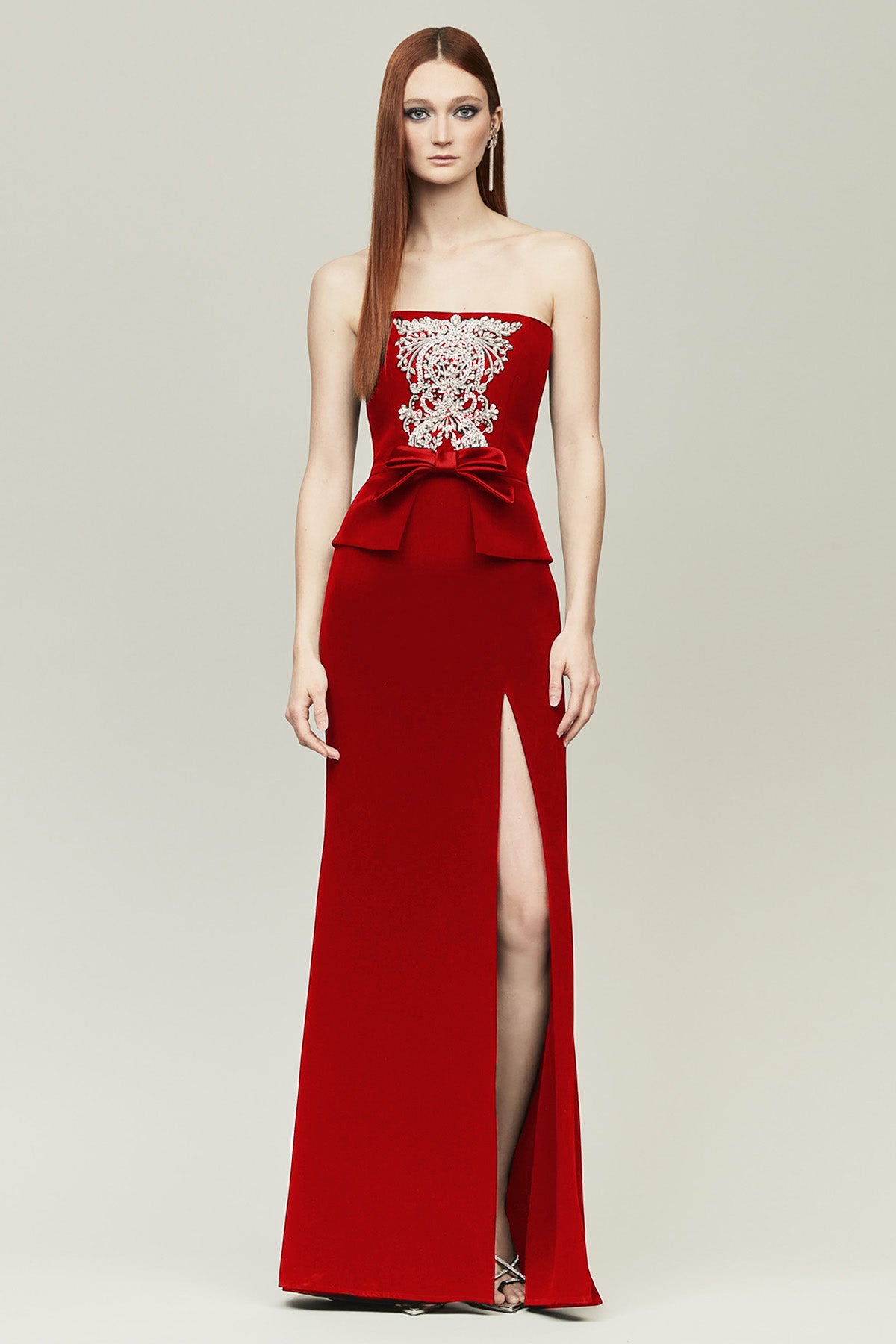 A woman modeling a strapless red evening gown with a high thigh slit and a large bow at the waist, featuring a white embroidered bodice, against a light gray studio background.