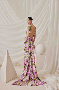 A woman stands with her back to the camera wearing a floor-length pink floral gown with a draped train and open back, posed in a studio with white draped fabric and geometric props.