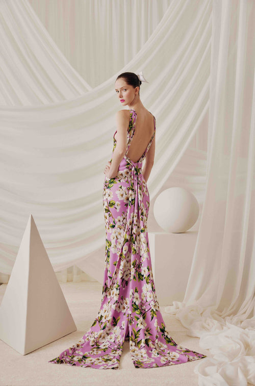 A woman stands with her back to the camera wearing a floor-length pink floral gown with a draped train and open back, posed in a studio with white draped fabric and geometric props.