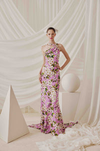 A model wears a lavender pink floral halter-neck evening gown with white and green cherry blossom motifs, posed in front of a draped white fabric backdrop with geometric white props.