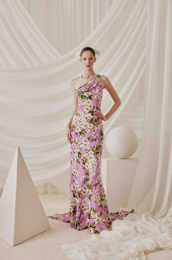 A model wears a lavender pink floral halter-neck evening gown with white and green cherry blossom motifs, posed in front of a draped white fabric backdrop with geometric white props.