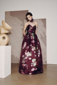 Woman in a floral evening gown standing in an artistic setting with sculptures.