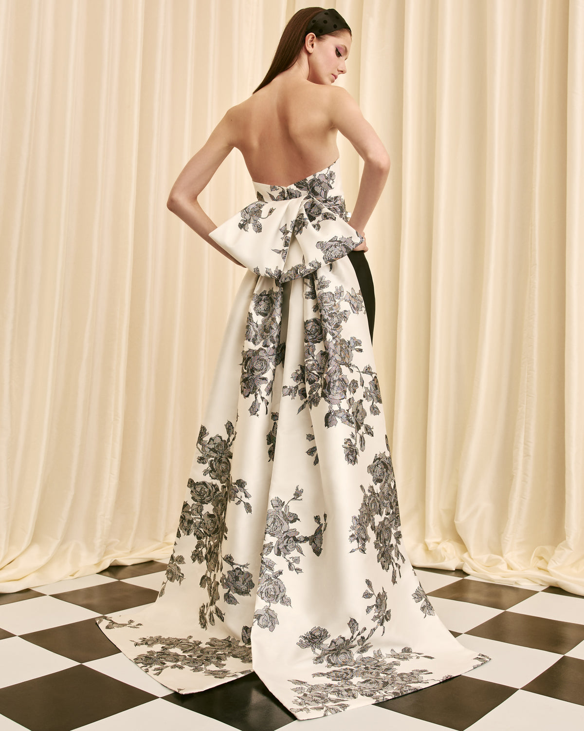 Woman wearing a floral evening gown on a checkered floor with curtains in the background.  Back view of the Dress.