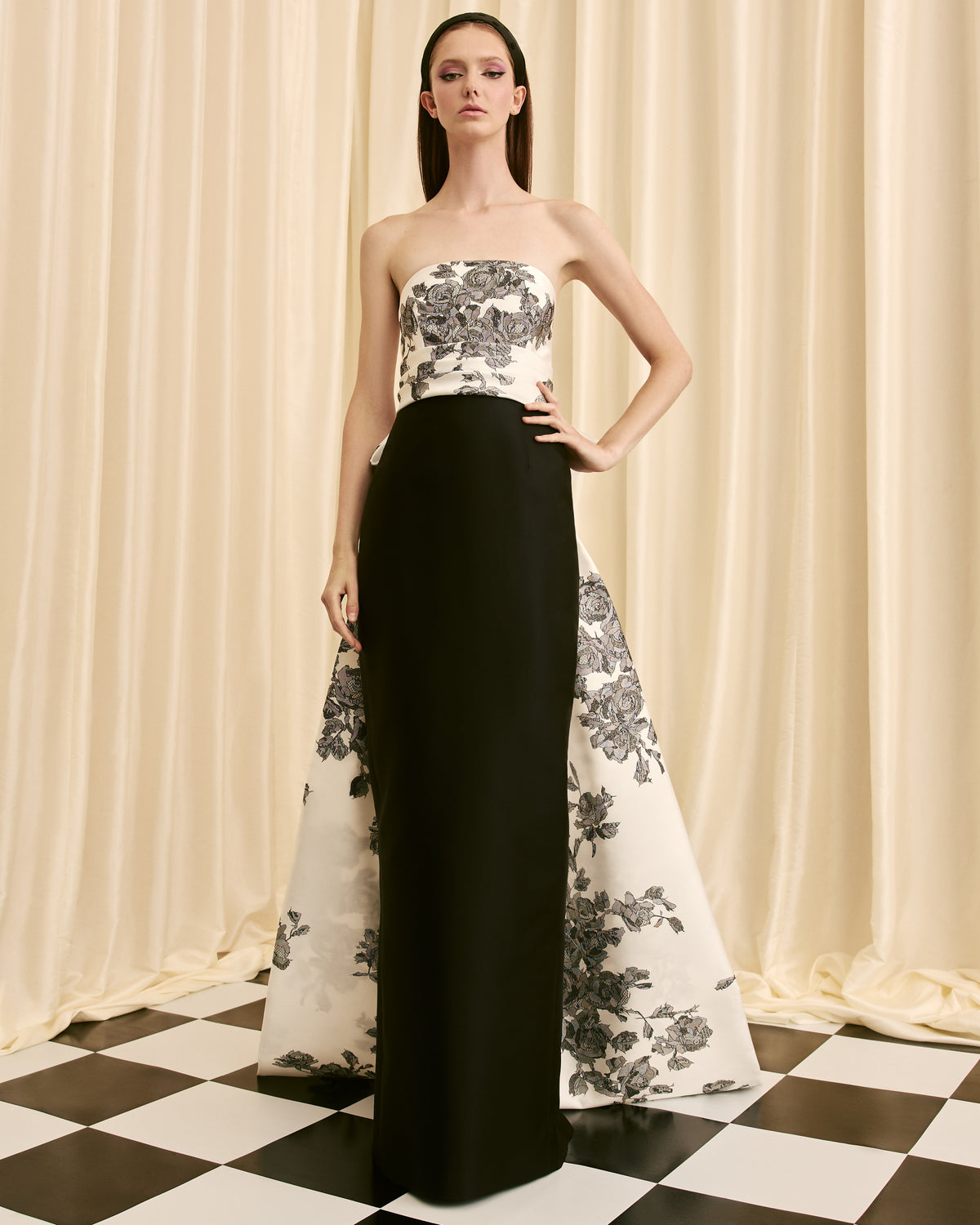 Model wearing a strapless floral top and black and white floral gown against a checkered floor and curtain background.