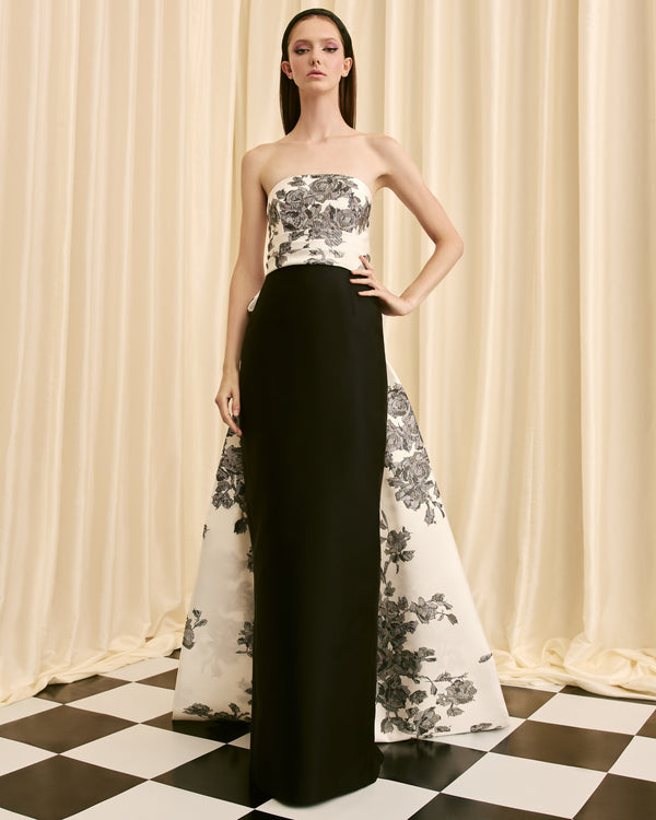 Model wearing a strapless floral top and black and white floral gown against a checkered floor and curtain background.