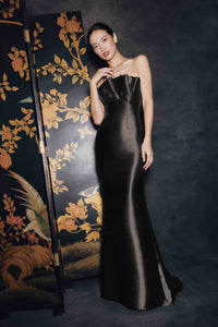 A woman models a strapless black satin mermaid gown with a structured bodice and a floor-length silhouette, posed next to a decorative black folding screen with gold floral and bird artwork against a dark studio backdrop.