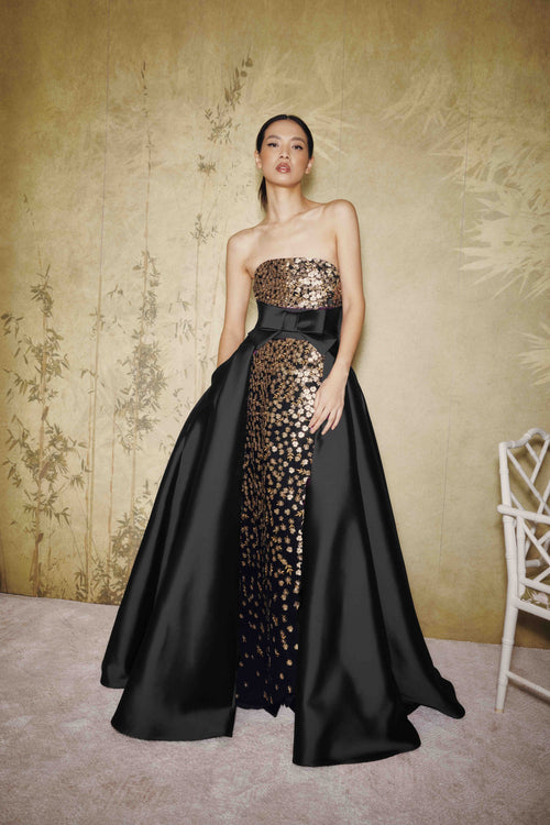A model wears a strapless black evening gown with a wide black overskirt and a central panel of gold sequined floral embroidery, standing against a textured beige wall.