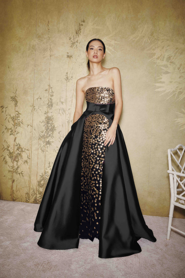 A model wears a strapless black evening gown with a wide black overskirt and a central panel of gold sequined floral embroidery, standing against a textured beige wall.