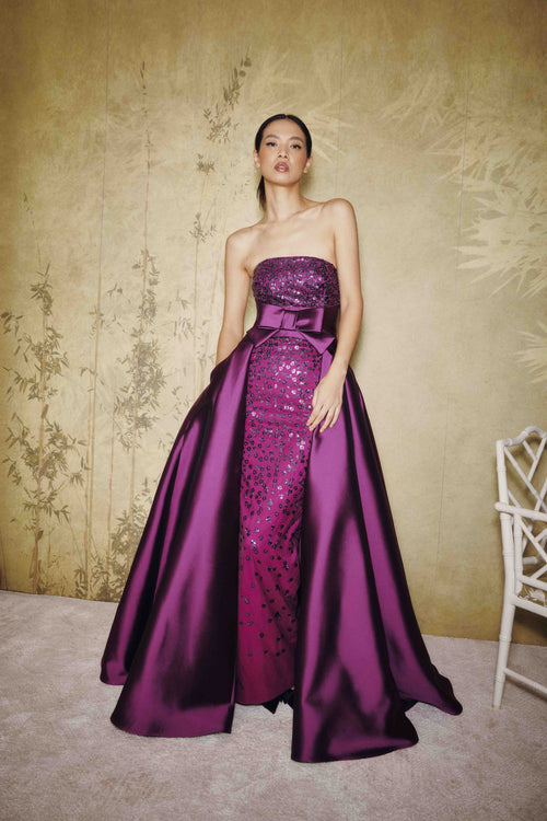 A woman models a strapless floor-length gown in rich purple. The dress features a sequined bodice and a satin front panel with a bow at the waist, set against a textured beige wall with subtle botanical patterns.