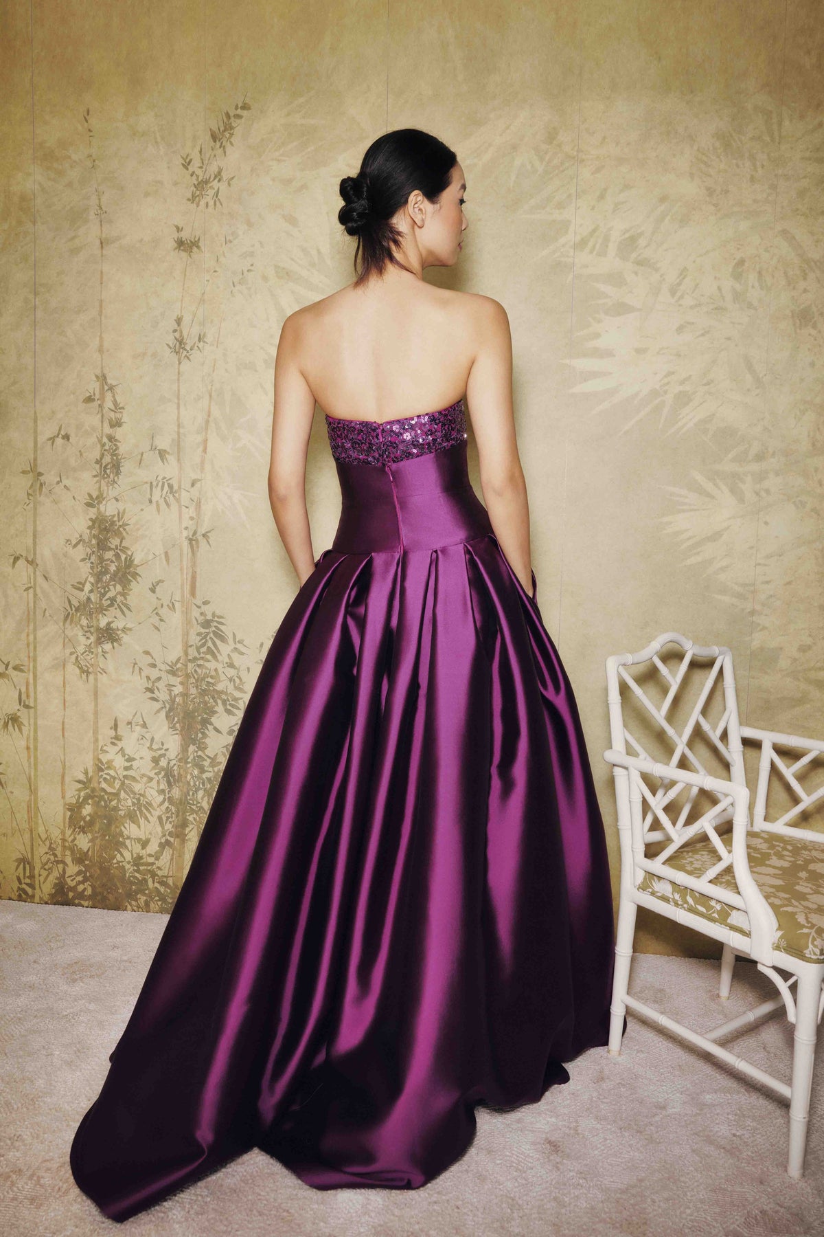 A woman stands with her back to the camera wearing a strapless ball gown in deep purple satin. The dress features a beaded waistband and a voluminous skirt.