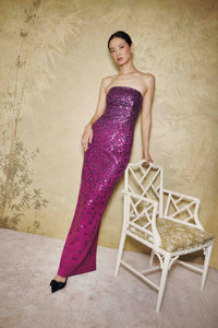 A woman models a strapless, floor-length magenta sequined gown next to a white decorative chair against a textured beige wall.