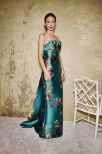 A woman stands in a studio wearing a strapless satin gown with a teal-green base and large floral print in pink, red, and green. The background is a textured beige wall with a white chair on the right.