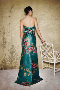 A woman stands with her back to the camera wearing a strapless satin gown in deep teal with a large bow at the waist and a floral print in pink, red, and green. She is posed in front of a textured beige wall next to a white chair.