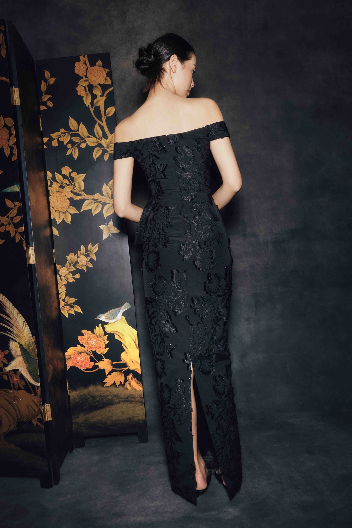 A woman stands with her back to the camera wearing an off-the-shoulder black lace evening gown with a thigh-high slit. She is posed in front of a dark textured backdrop and a decorative black folding screen with gold floral and bird artwork.