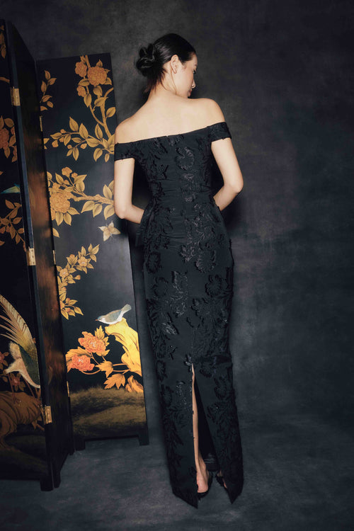 A woman stands with her back to the camera wearing an off-the-shoulder black lace evening gown with a thigh-high slit. She is posed in front of a dark textured backdrop and a decorative black folding screen with gold floral and bird artwork.