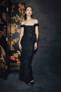 A woman stands in a dark studio setting wearing an off-the-shoulder black lace gown with a fitted waist and flared skirt. A dark floral folding screen is visible to her left.