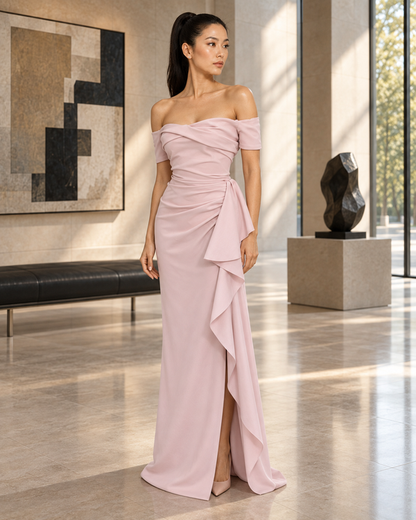 A woman models an off-the-shoulder floor-length gown in a modern, sunlit interior with large windows, abstract wall art, and a sculpture. The dress is a soft blush pink with a draped, layered ruffle design along the skirt and waist.