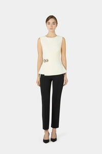 A model stands against a light gray studio background wearing a sleeveless cream peplum top with a decorative gold-tone waist detail and black cropped trousers with black heels.