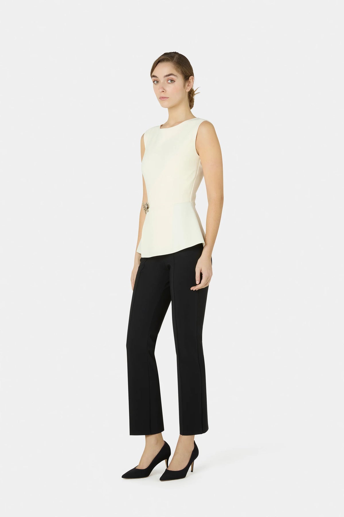 A woman modeling a sleeveless cream top and black cropped trousers with black heels, standing against a light gray studio background.
