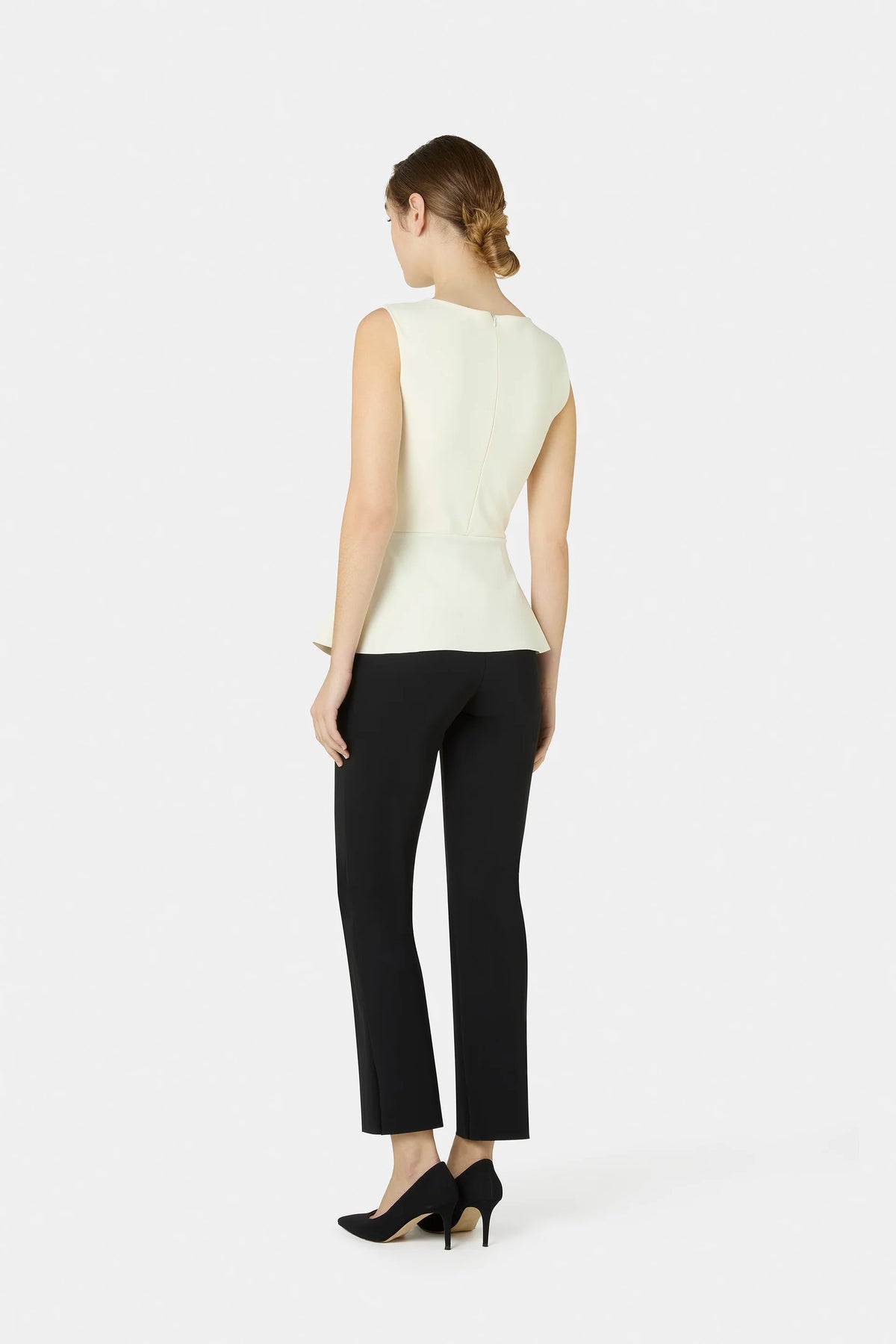 A woman shown from the back wearing a sleeveless cream peplum top, black cropped trousers, and black heels against a light gray studio background.