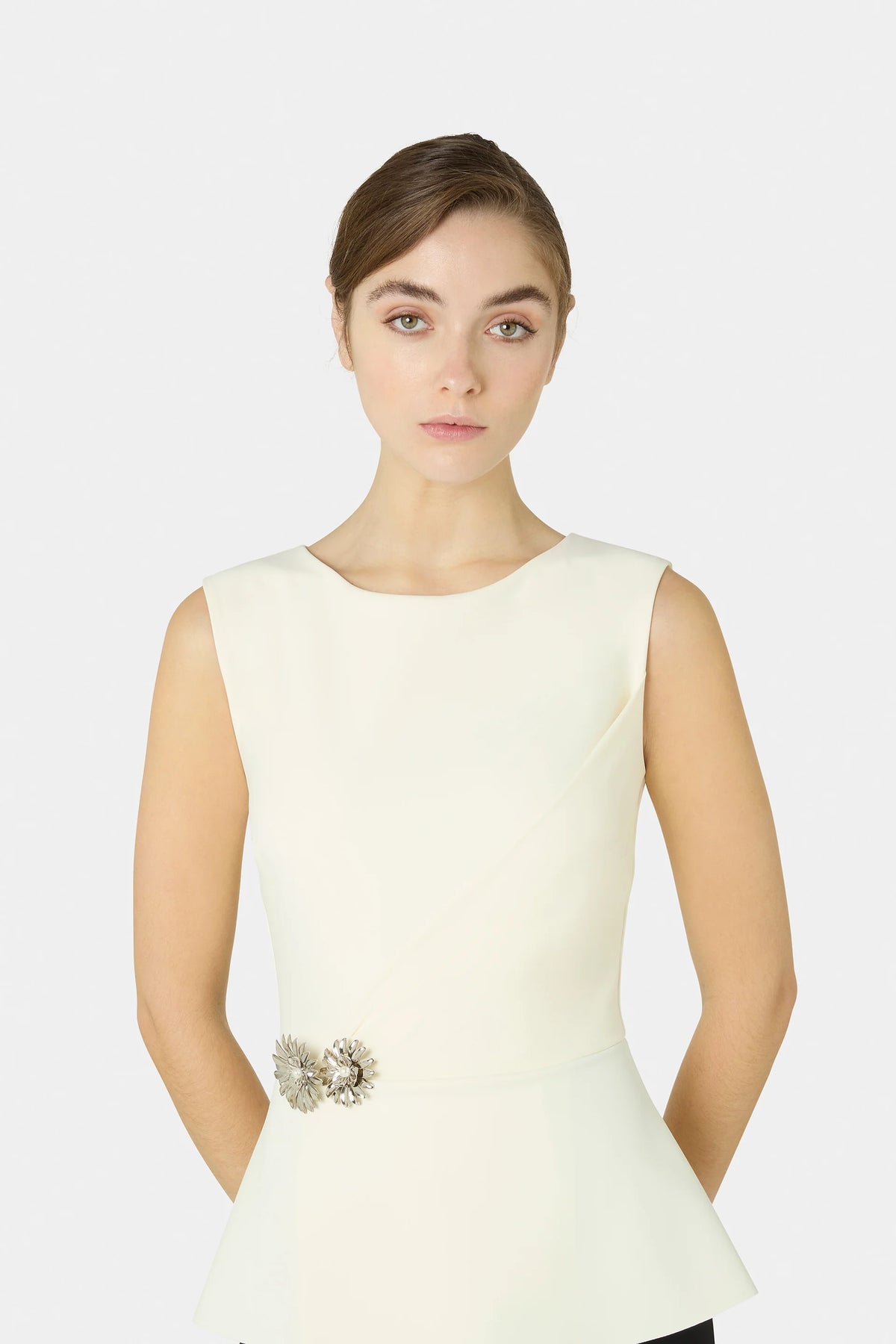 A woman modeling a sleeveless cream-colored peplum top with a decorative jeweled waist embellishment, set against a plain light gray studio background.