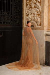 Model wearing Chic & Holland HF110562 gown with intricate beading and flowing cape at a formal couture setting.