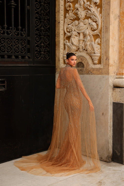 Model wearing Chic & Holland HF110562 gown with intricate beading and flowing cape at a formal couture setting.
