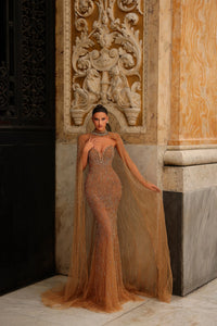 Model wearing Chic & Holland HF110562 gown with intricate beading and flowing cape at a formal couture setting.