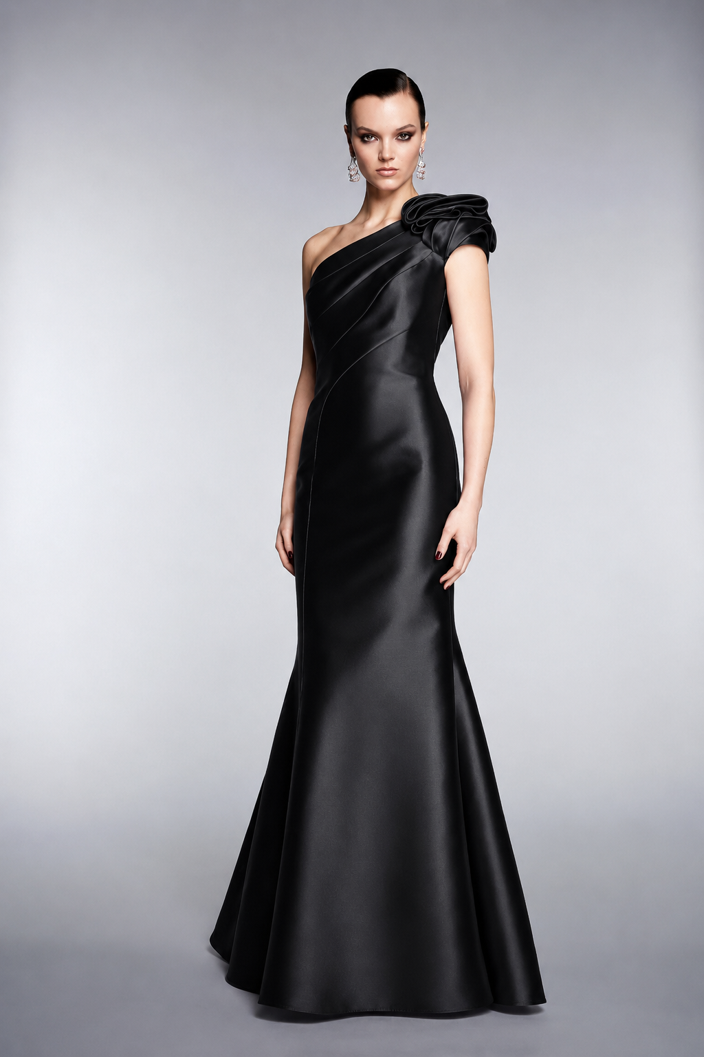 A model wearing a floor-length black satin one-shoulder evening gown with a dramatic ruffled shoulder detail, photographed against a neutral gray studio background.