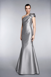 A woman modeling a floor-length one-shoulder evening gown in a smooth silver satin fabric, photographed against a neutral light gray studio background.