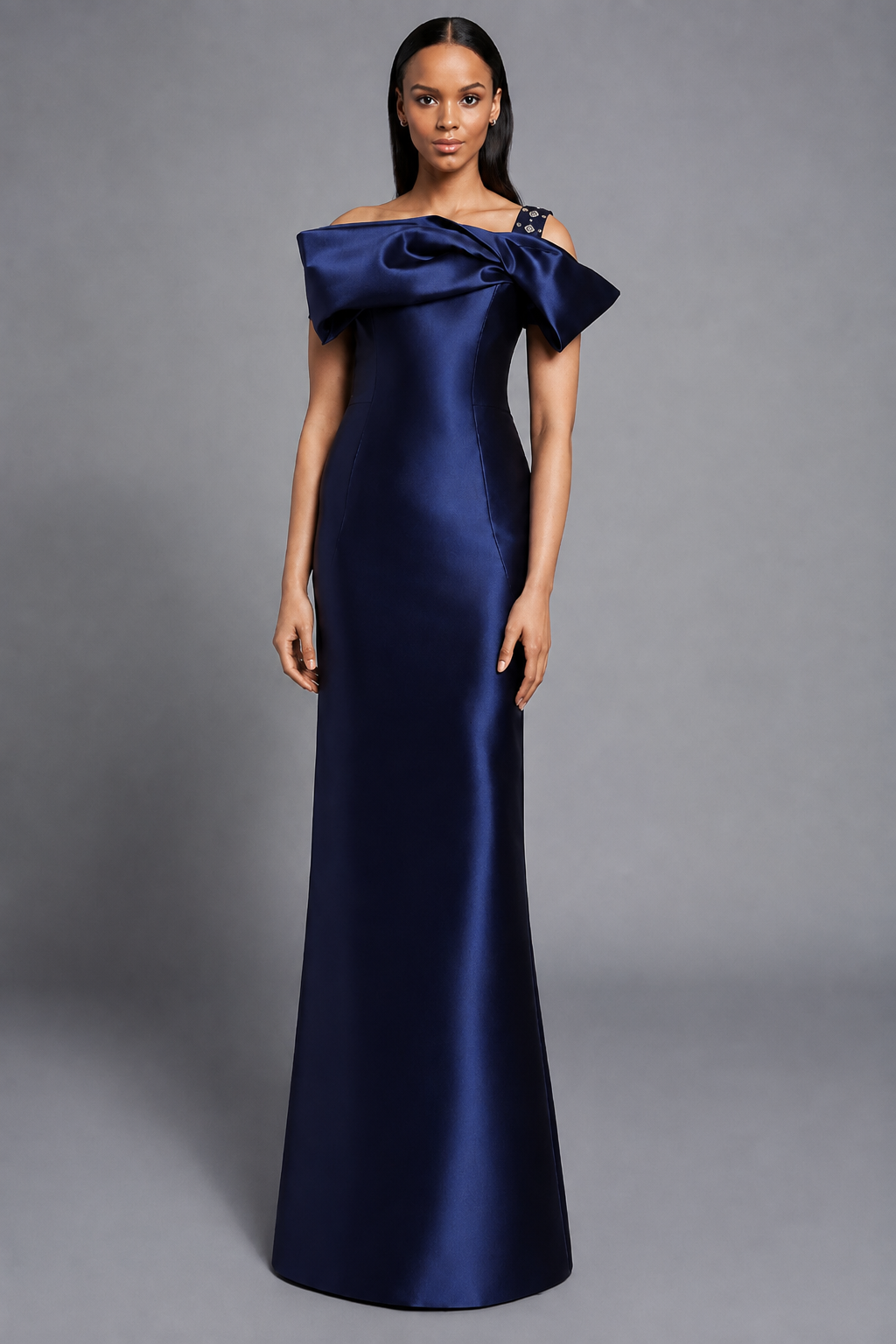 A woman modeling a floor-length off-the-shoulder gown in navy against a neutral gray studio background.