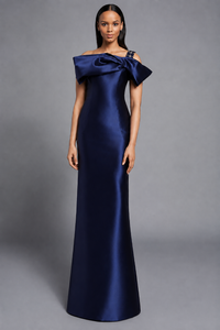 A woman modeling a floor-length off-the-shoulder gown in navy against a neutral gray studio background.