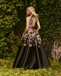 Woman in a black evening gown with floral embellishments standing against a green ivy wall.