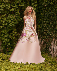 A woman stands outdoors in front of a dense green ivy wall, wearing a sleeveless halter-neck ball gown with a pale blush pink skirt and a floral bodice in pink, purple, and beige tones.