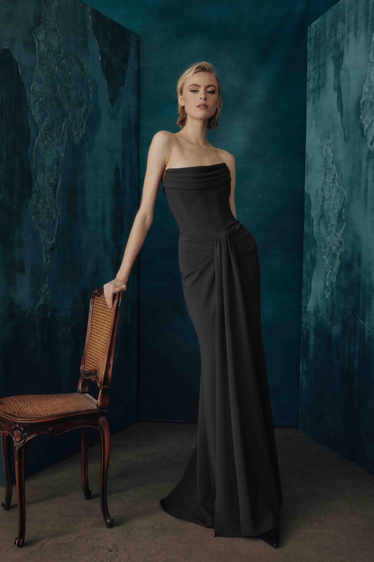 A woman models a strapless black evening gown in a moody studio setting with textured teal walls and a wooden chair beside her.