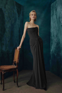 A woman models a strapless black evening gown in a moody studio setting with textured teal walls and a wooden chair beside her.