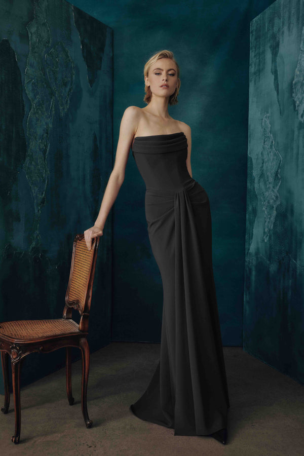 A woman models a strapless black evening gown in a moody studio setting with textured teal walls and a wooden chair beside her.