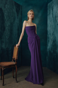 A woman models a strapless floor-length boysenberry gown in a dark teal studio setting, leaning against a wooden chair.