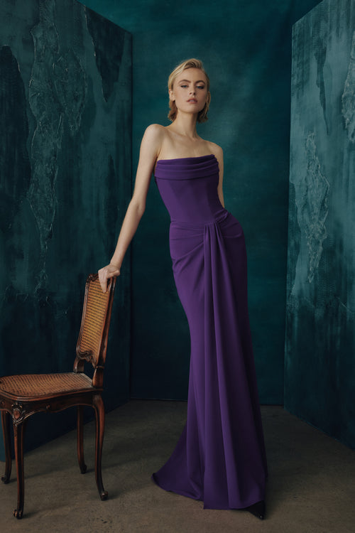 A woman models a strapless floor-length boysenberry gown in a dark teal studio setting, leaning against a wooden chair.