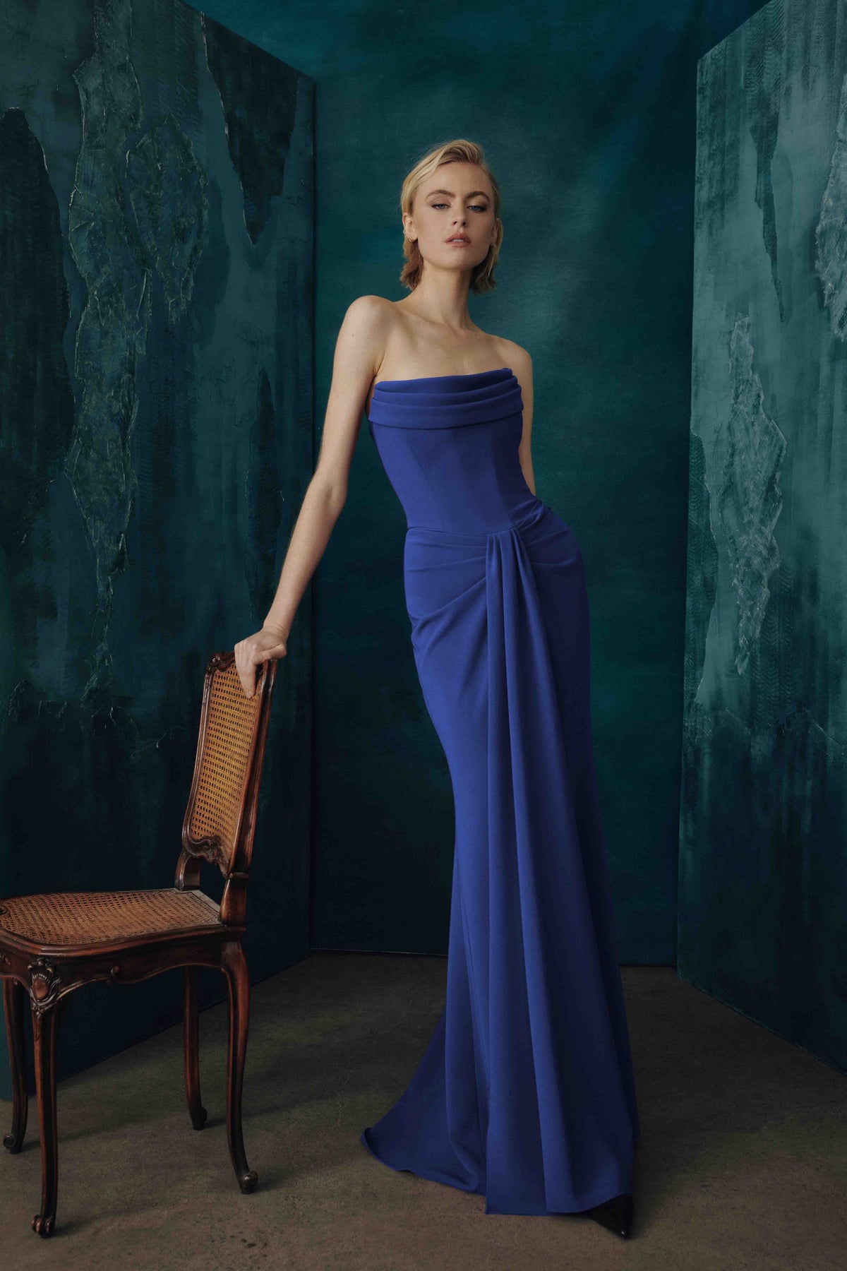 A woman models a strapless royal blue floor-length gown in a moody studio setting with dark teal textured walls and a wooden chair beside her.